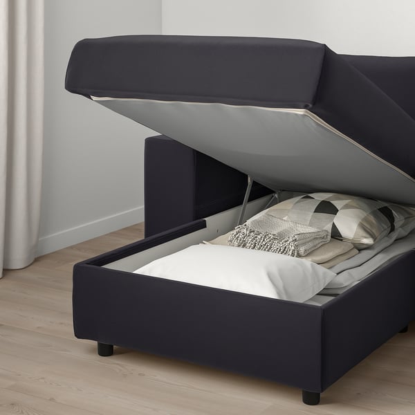 A grey upholstered sofa bed with a lift-up seat revealing a storage compartment and blanket.