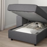 A grey upholstered sofa with storage underneath, partially opened to reveal blankets and pillows inside.