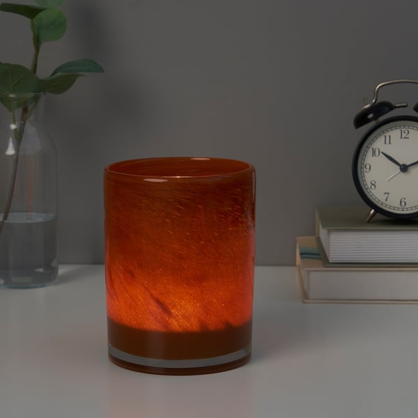 Orange candle holder with plant, books, clock, warm glow.
