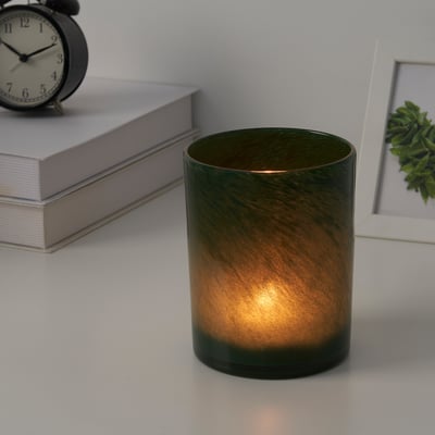 Cylindrical glass candle holder, lit, on white desk near books, clock, and plant.