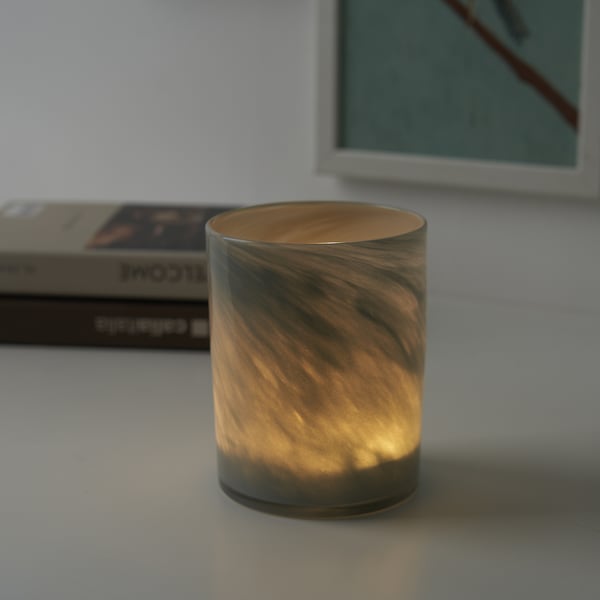 A lit candle in a textured glass holder sits on a white surface. The holders design reflects the warm glow, creating a cozy ambiance.