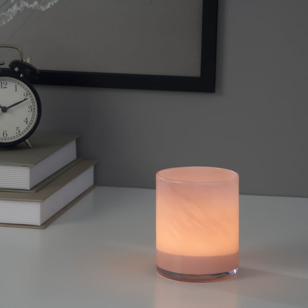A lit candle in a pink glass holder sits on a table beside books and a clock, creating a cozy atmosphere.