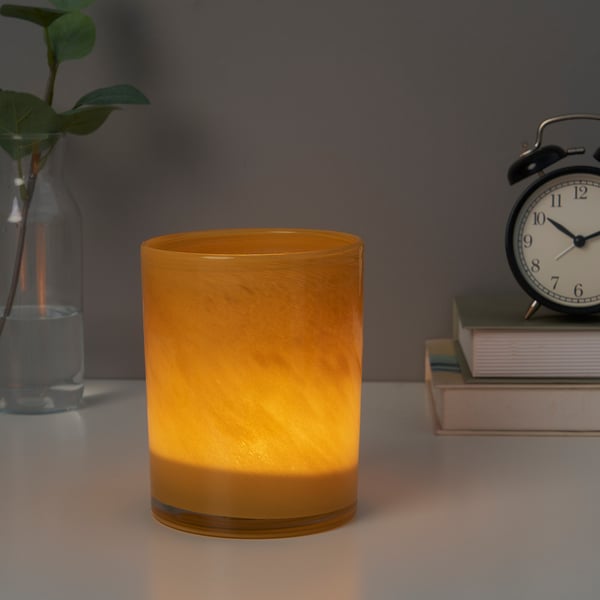 Orange glass candle holder, lit from within, on table beside clock and plant.