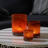 Three glowing amber glass candle holders of varying sizes on a white table. They emit a warm light and cast decorative shadows.