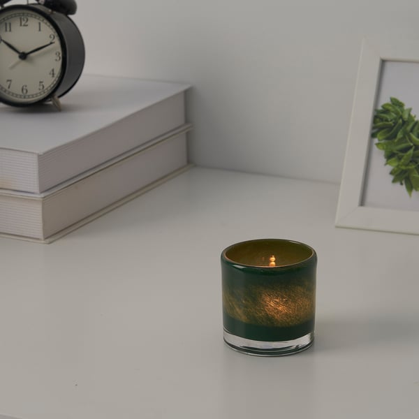 Candle in green glass holder on white surface with books, clock, and framed plant.