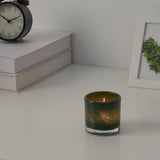 Candle in green glass holder on white surface with books, clock, and framed plant.