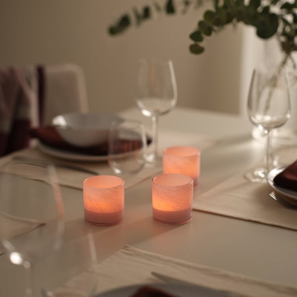 Table set with glowing VINDSTILLA pink tealight holders, clear glasses, plates, cutlery, and foliage. Holders small compared to other items.
