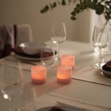 Table set with glowing VINDSTILLA pink tealight holders, clear glasses, plates, cutlery, and foliage. Holders small compared to other items.