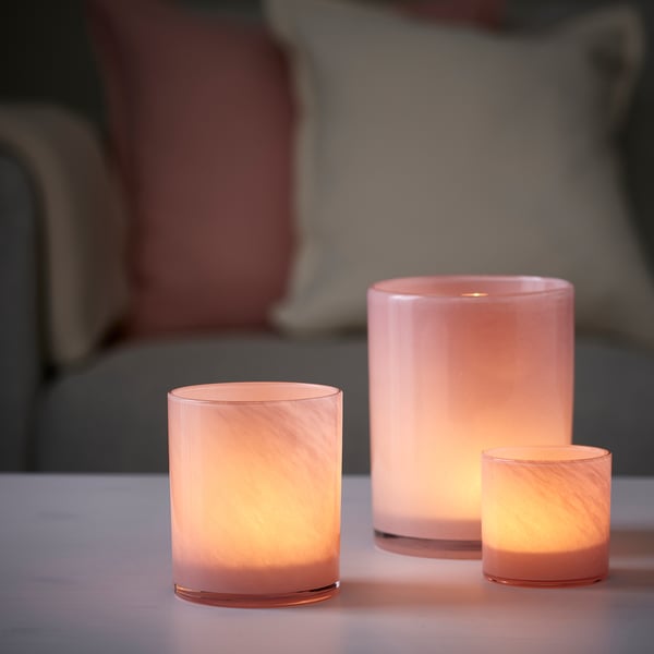 Three pink glass VINDSTILLA tealight holders of varying sizes, mouth-blown with a decorative pattern, glowing warmly on a table.