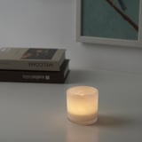 Lit candle in glass holder on table, warm glow, with books and framed picture.