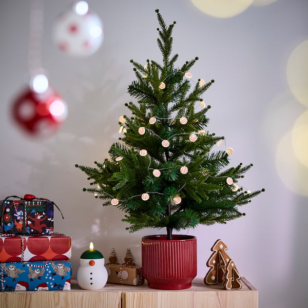 Christmas tree in red pot, adorned with white ornaments and lights, on wooden shelf with gifts and decorations.