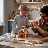 Man & child decorate gingerbread house with icing, surrounded by candy and bowls on a wooden table.