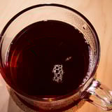 Clear glass mug filled with dark, bubbly, spiced beverage. Visible vapors suggest warmth. Resting on a wooden surface.