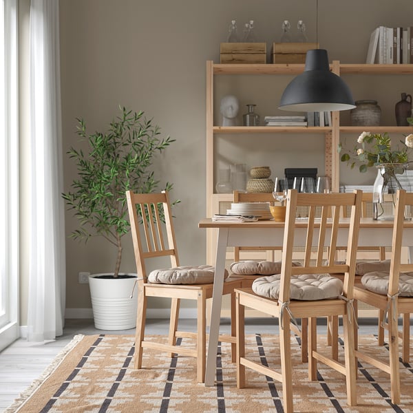 Dining area with wooden chairs & beige cushions, rug, plant, & shelves with decor.