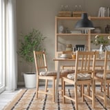 Dining area with wooden chairs & beige cushions, rug, plant, & shelves with decor.