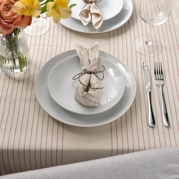 Table set with white plates, folded napkins, silverware, wine glass, and striped tablecloth; flowers in vase.