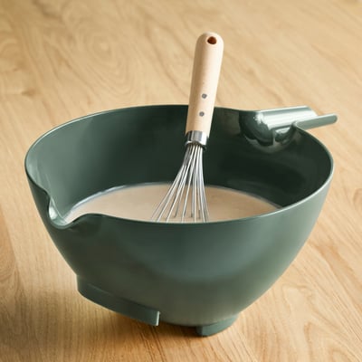 Dark green mixing bowl with a wooden handle used for whisking, resting on a wooden surface.