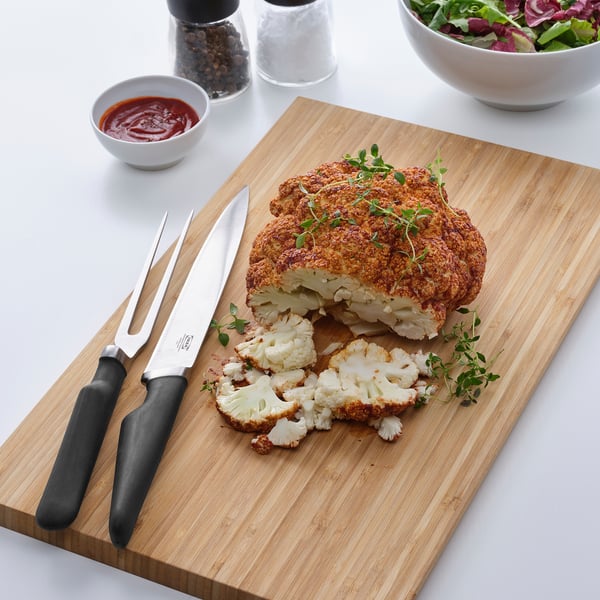 A wooden cutting board displays a carved roasted cauliflower with a carving knife and fork. A small bowl of ketchup and a pepper mill are nearby.