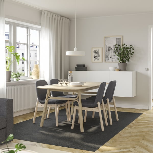 Modern dining set with grey chairs and light wood table. Minimalist decor with plant and rug.