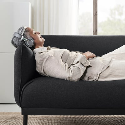 En äldre person kopplar av i en grå ÄPPLARYD-soffa, iförd hörlurar, och njuter av en lugn stund med slutna ögon.