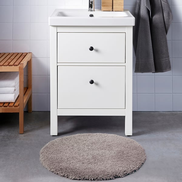 En vit kommod med handfat och lådor, med en plysch, beige ALMTJÄRN badrumsmatta och en träbänk med handdukar och en korg.