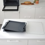 Modern köksbänk med tre staplade svarta bakplåtar bredvid en öppen ugn och kylgaller.