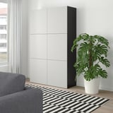 Stora, moderna vita BESTÅ-skåp tornar upp sig i vardagsrummet, höga bredvid grå soffa och stor inomhusväxt, med elegant, minimal design.