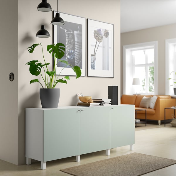 Ljusblått BESTÅ sideboard med 3 dörrar, vita ben och en stor växt på toppen. Den ligger bredvid en orange soffa i ett modernt vardagsrum.
