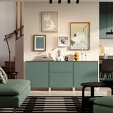 Modern vardagsrum med teal BESTÅ-skänk , konstvägg, grön soffa och randig matta under trappan.