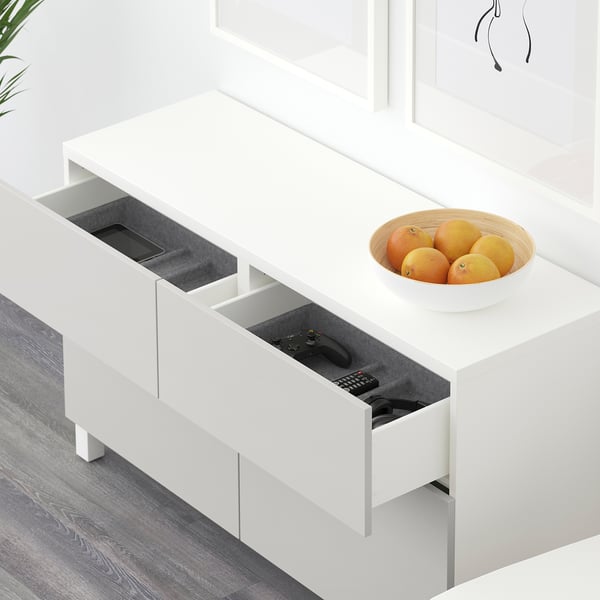 Vitt BESTÅ-sideboard med öppna lådor som avslöjar en telefon, spelkontroll och fjärrkontroll i en låda och apelsiner i en skål ovanpå.