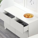Vitt BESTÅ-sideboard med öppna lådor som avslöjar en telefon, spelkontroll och fjärrkontroll i en låda och apelsiner i en skål ovanpå.