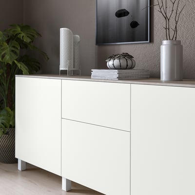 Minimalistiskt rum med vitt skåp, ljusgrå topppanel, staplade böcker, växter och väggkonst.