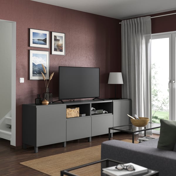 Modernt vardagsrum med mörkröda väggar, med en grå BESTÅ-tv-enhet, en platt-tv, elegant inredning och stora fönster med utsikt över grönska.
