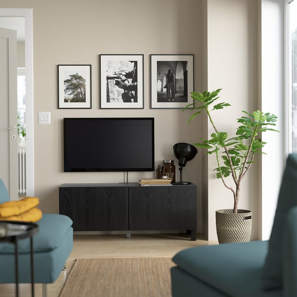 Modernt vardagsrum med tv-bänken BESTÅ, plattskärm, blågröna stolar, svart lampa, gul pläd, växt och svartvita ramar.