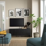 Modernt vardagsrum med tv-bänken BESTÅ, plattskärm, blågröna stolar, svart lampa, gul pläd, växt och svartvita ramar.
