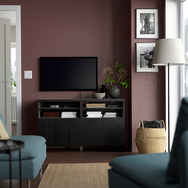 Modernt vardagsrum med tv-bänken BESTÅ, böcker, vaser och plattskärms-tv mot mörklila vägg. Blå och svarta möbler, stor lampskärm.