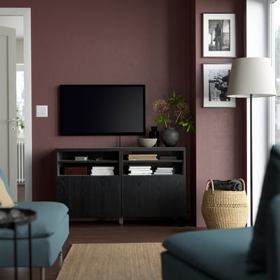 Modernt vardagsrum med tv-bänken BESTÅ, böcker, vaser och plattskärms-tv mot mörklila vägg. Blå och svarta möbler, stor lampskärm.