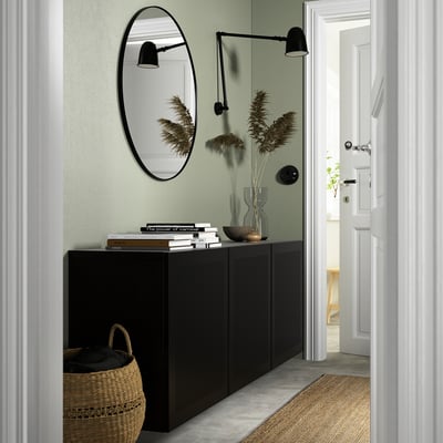 Modern hall med svart BESTÅ-enhet, spegel och lampor. Blomsterarrangemang, böcker, skål och korg.
