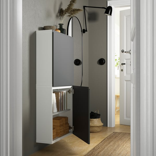 Öppet förvaringsskåp med hyllor, inklusive flätad korg och tidningar, monterade på en grå vägg. Svart vägglampa och vävd korg i närheten.