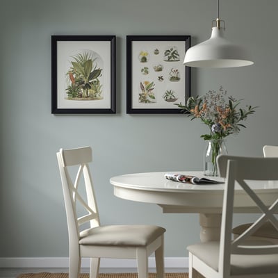 Två svarta inramade växtkonsttryck ovanför vitt matbord med blommor, hängande ljus, ljusblå vägg.