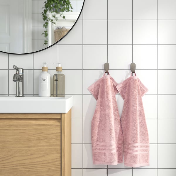 BROKGLIM rosa handdukar hänger snyggt på kromkrokar bredvid ett modernt handfat med vit bänkskiva, spegel och vedskåp. Mjuk, absorberande och ekologisk.