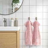 BROKGLIM rosa handdukar hänger snyggt på kromkrokar bredvid ett modernt handfat med vit bänkskiva, spegel och vedskåp. Mjuk, absorberande och ekologisk.