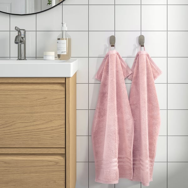 Badrumsscen med ljusrosa BROKGLIM handdukar som hänger, matchande vitt handfat, träskåp och modern inredning. Elegant och organiserat utseende.