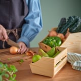 En person skär lök på en skärbräda av trä, med andra grönsaker i trälådor på ett bord.