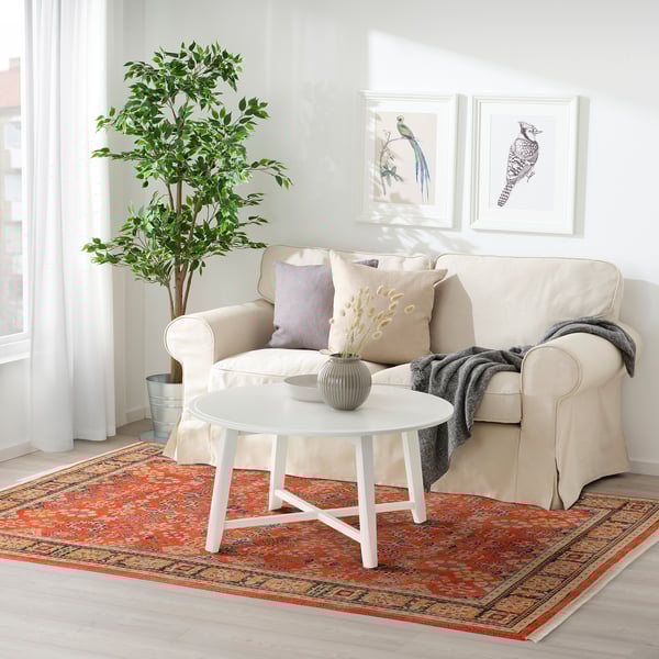 Minimalistiskt vardagsrum med beige soffa, vitt soffbord och röd matta med blommiga mönster.
