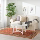 Minimalistiskt vardagsrum med beige soffa, vitt soffbord och röd matta med blommiga mönster.