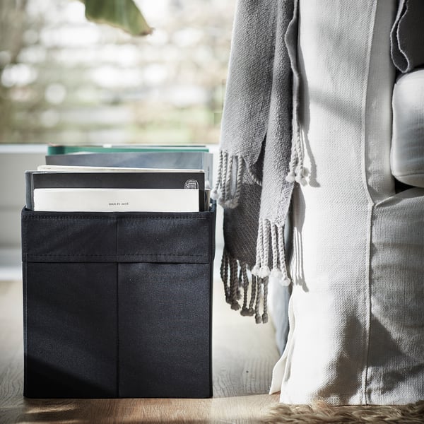 Svart tygförvaringslåda nära en grå soffa, som rymmer böcker med en grå och vit filt draperad i närheten.