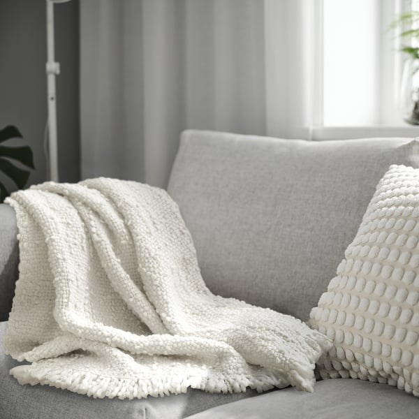 En vit DYSTERMAL pläd på en grå soffa. Filten är strukturerad, har fransar och ser mysig ut. Den är större än kudden som den täcker delvis.