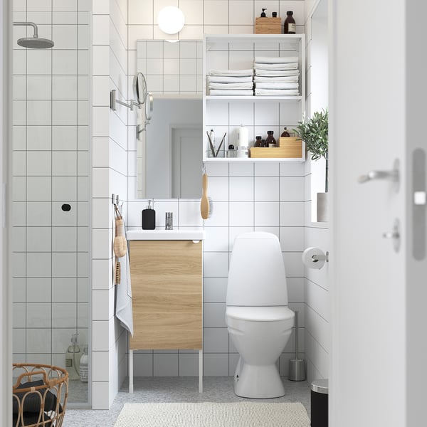 Föreställer ett modernt badrum med vitt kakel, ett träskåp och en elegant toalett. Badrummet har en dusch, spegel, hyllor med handdukar och en växt.