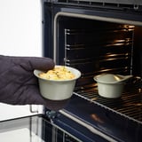 En hand som håller en liten keramisk form med ugnsbakad mat, redo att placeras i en öppen ugn, bredvid en annan liknande form inuti.